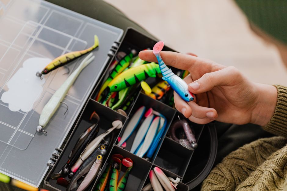 Person holding a colorful fishing lure above a well-organized tackle box full of assorted baits.